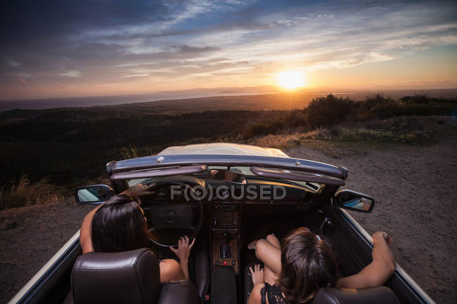 Dos mujeres jóvenes en coche convertible, conduciendo a lo largo de la carretera panorámica, vista trasera - foto de stock