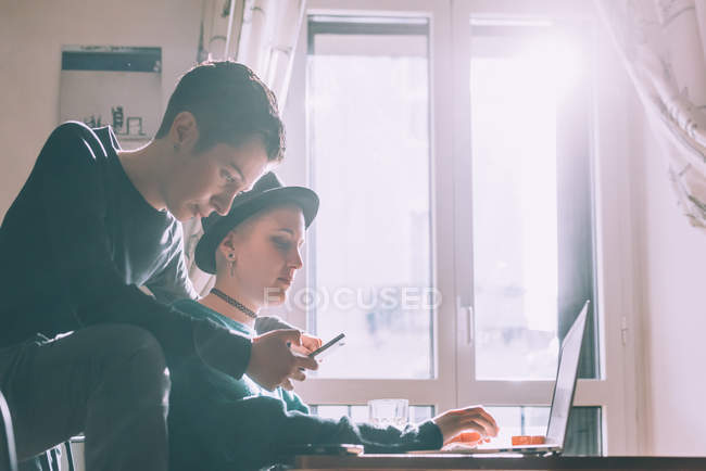 Dos amigas jóvenes usando smartphone y laptop en la sala de estar - foto de stock