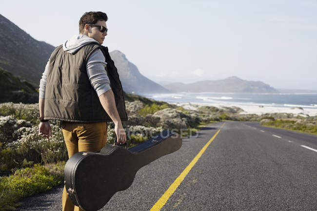 Vue arrière d'un jeune homme marchant sur une route côtière portant un étui à guitare, Cape Town, Western Cape, Afrique du Sud — Photo de stock