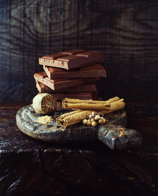 Pau de canela, noz-moscada e barras de chocolate quebradas empilhadas em placa de corte de madeira — Fotografia de Stock