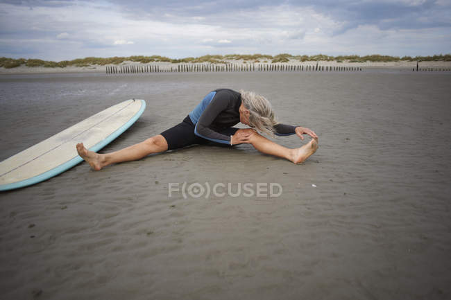 Seniorin sitzt auf Sand, dehnt sich, Surfbrett neben ihr — Stockfoto