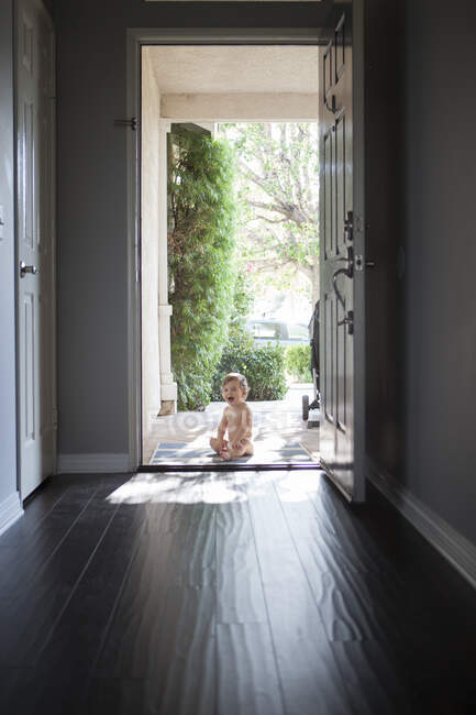 Vue à travers le hall d'entrée du bébé garçon assis à l'extérieur à la porte d'entrée ouverte, regardant la caméra ouverte bouche ouverte — Photo de stock