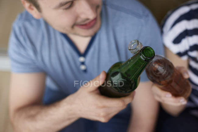 Jovem casal sentado ao ar livre, brindando com cervejas engarrafadas — Fotografia de Stock