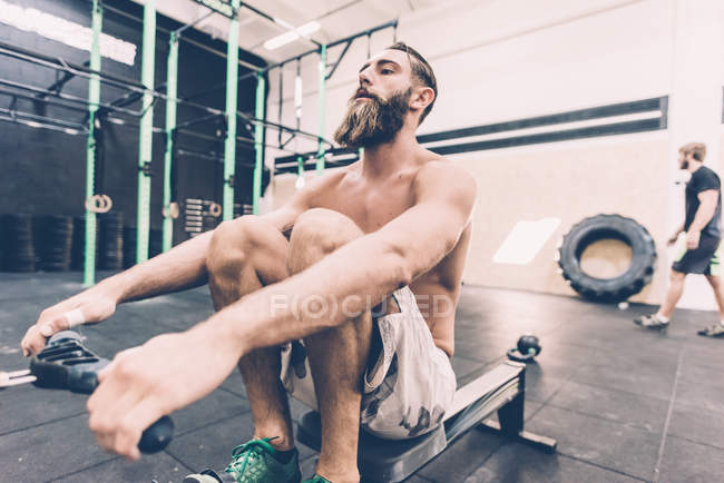 Entrenamiento de entrenador cruzado masculino joven en máquina de remo en el gimnasio - foto de stock