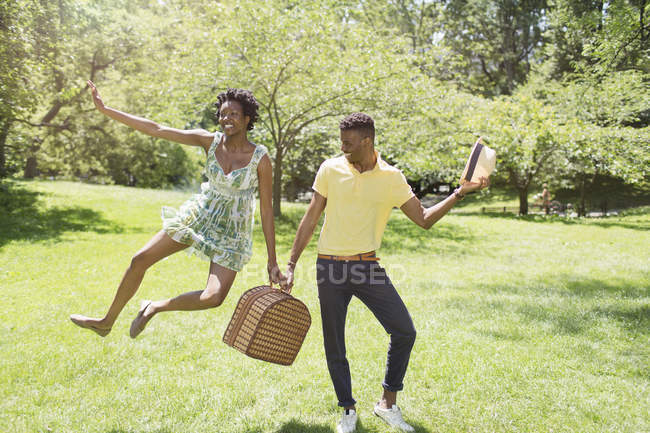 Casal jovem com cesta de piquenique no parque, com salto feminino no ar — Fotografia de Stock