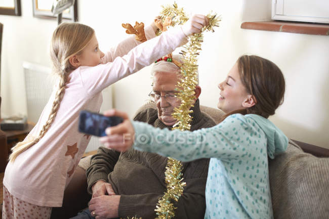 Sisters taking smartphone selfie while putting tinsel on sleeping grandfather — Stock Photo