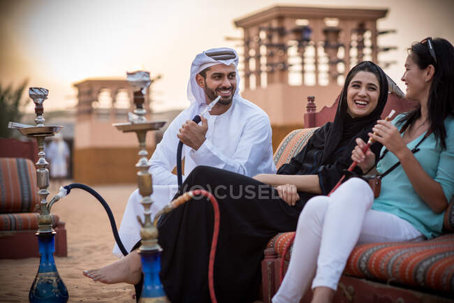 Local couple wearing traditional clothes smoking shisha on sofa with female tourist, Dubai, United Arab Emirates — Stock Photo