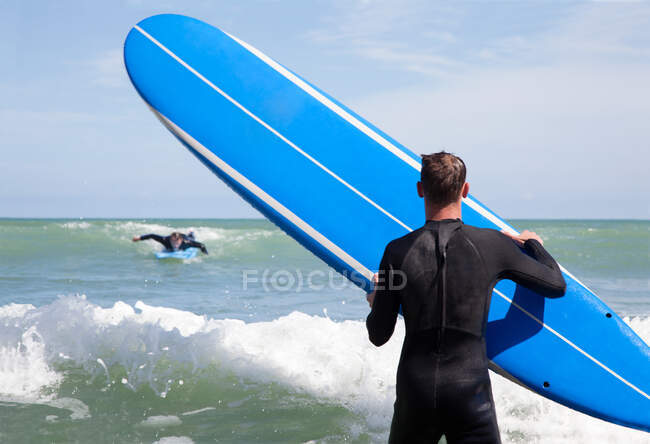 Vista trasera del surfista masculino con tabla de surf viendo amigo surf - foto de stock