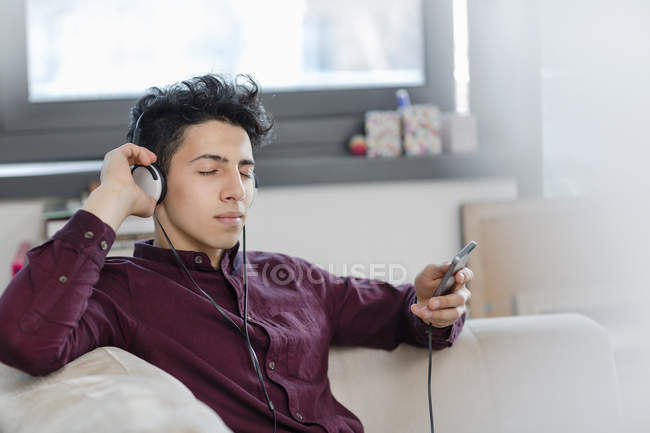 Jovem no sofá usando fones de ouvido segurando smartphone — Fotografia de Stock