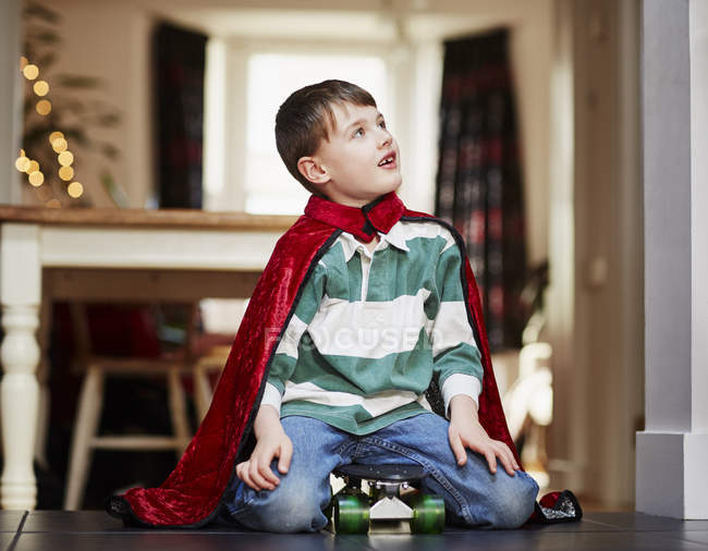 Jeune garçon assis sur une planche à roulettes portant une cape — Photo de stock