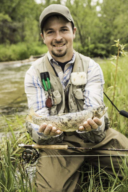 Hombre arrodillado en la orilla del río sosteniendo peces recién capturados mirando a la cámara sonriendo - foto de stock
