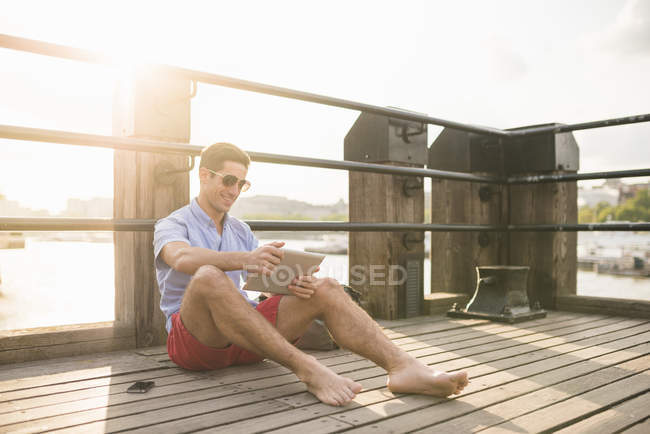 Молодой человек сидит на пирсе с помощью цифрового планшета — стоковое фото