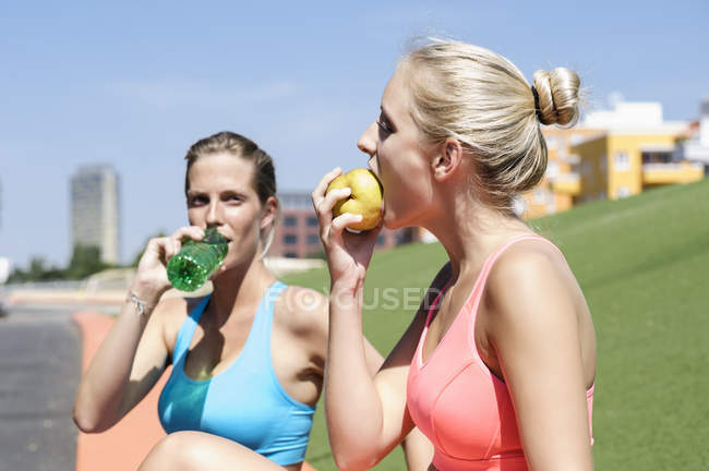 Молодые спортсменки берут перерыв на питание, едят фрукты и питьевую воду из бутылки на лугу — стоковое фото