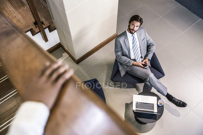 Vista de ángulo alto del hombre de negocios en el lobby sosteniendo el teléfono inteligente mirando hacia arriba - foto de stock