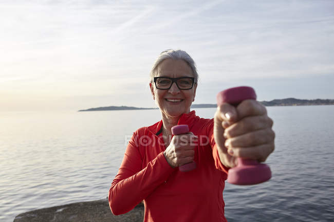 Mature woman beside water, exercising with hand weights, smiling — Stock Photo