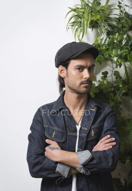 Retrato de jovem hipster masculino usando tampa plana em pé na frente da parede da folhagem — Fotografia de Stock