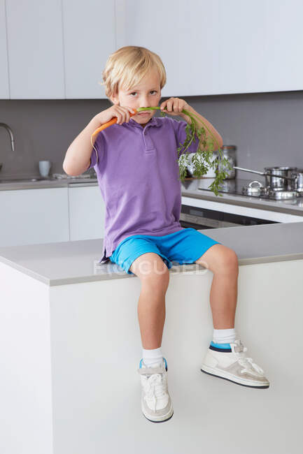 Boy sitting on side in kitchen with carrot moustache — Stock Photo