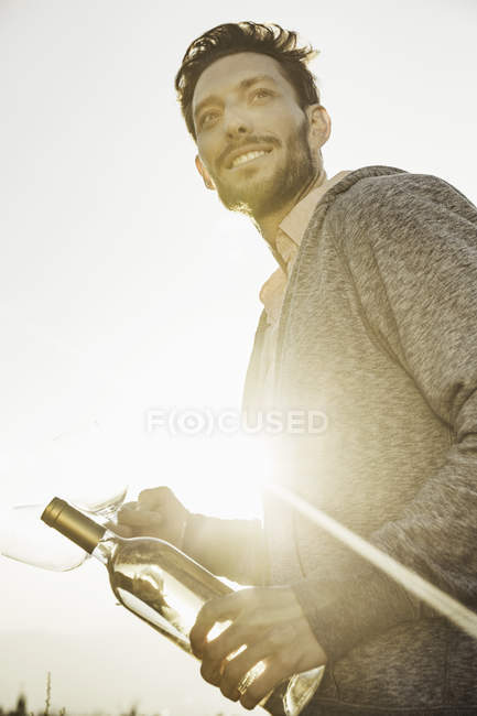 Homem no campo segurando garrafa de vinho e copos de vinho olhando para a câmera sorrindo — Fotografia de Stock