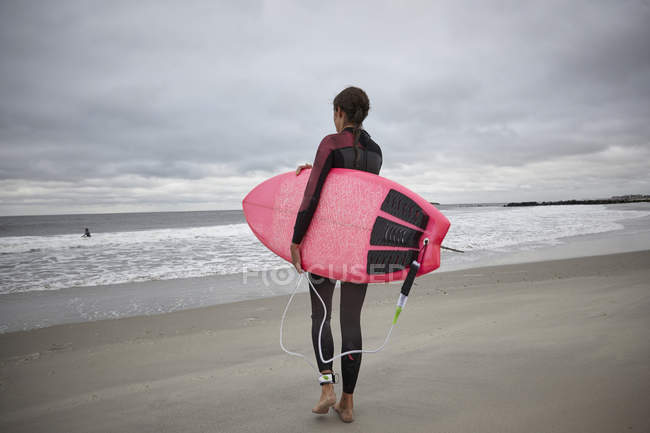 Вид сзади на женщину-серфингистку, несущую доску для серфинга в море на Рокавей Бич, Нью-Йорк, США — стоковое фото