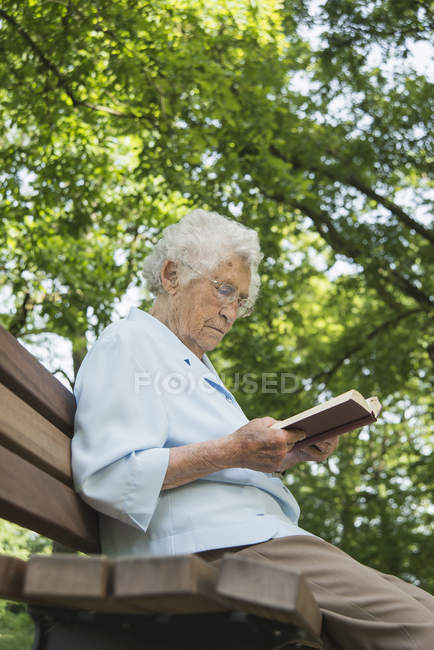 Старшая женщина, сидящая на скамейке в парке, читает Библию — стоковое фото