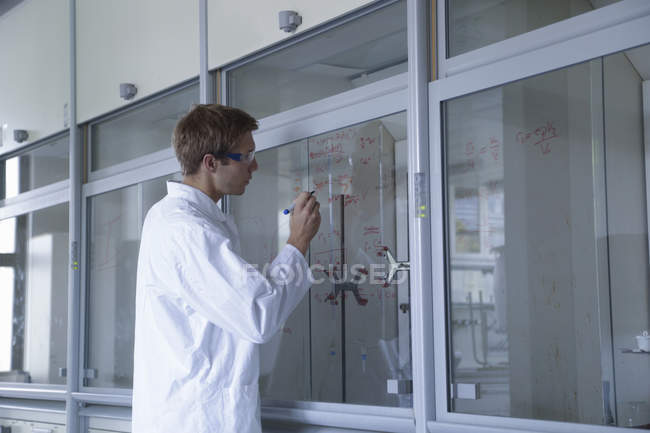 Joven científico masculino escribiendo resultados en la ventana del armario de humo - foto de stock