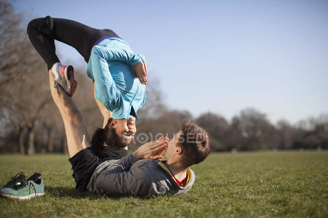 Молодая женщина балансирует на вершине человека практикуя йогу позы в парке — стоковое фото