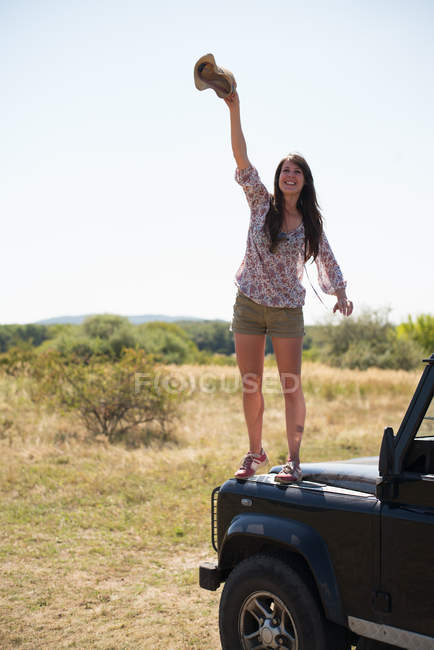Mid adult woman standing on hood of car waving hat — Stock Photo