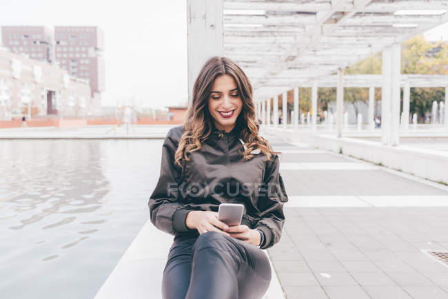 Mujer joven sentada al aire libre, usando smartphone, sonriendo - foto de stock