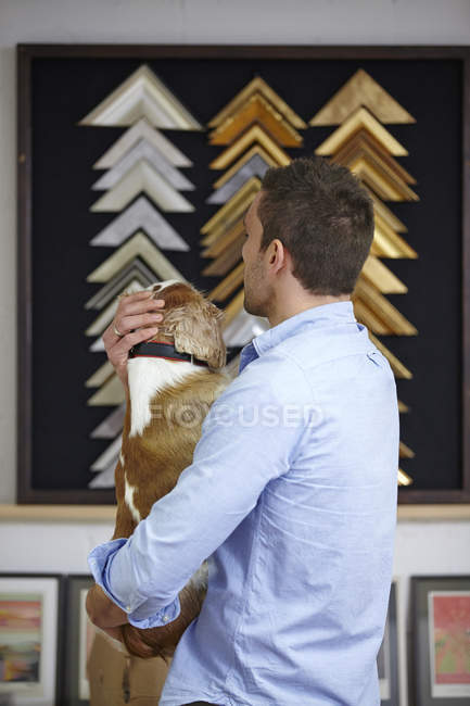 Man carrying dog whilst choosing frame in picture framers workshop — Stock Photo