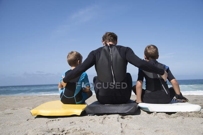 Rear view of man and two sons sitting on bodyboards, Laguna Beach, California, USA — Stock Photo