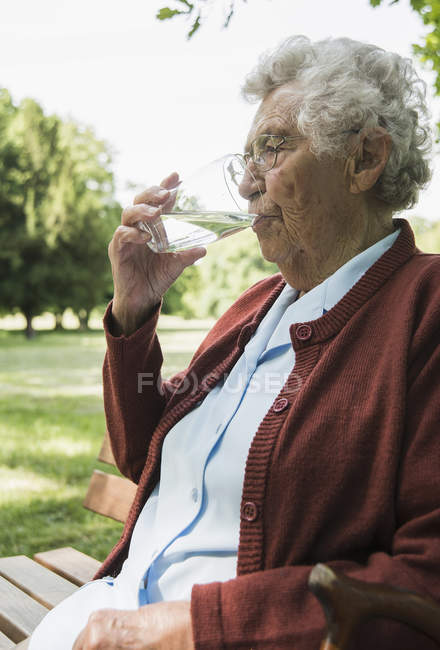 Seniorin sitzt auf Parkbank und trinkt Glas Wasser — Stockfoto