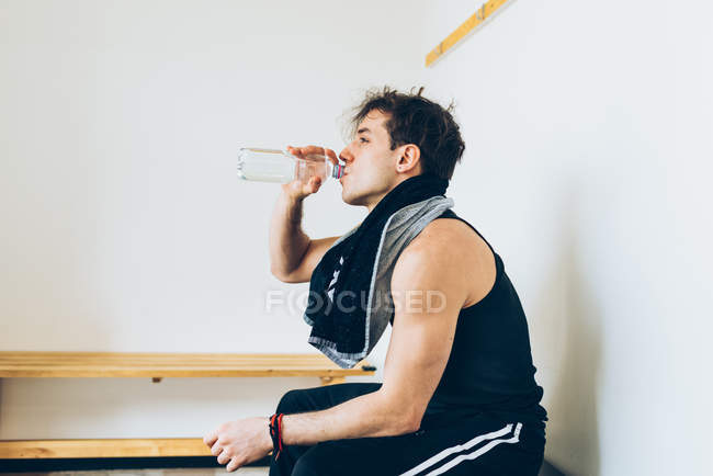 Мужчина сидит в раздевалке и пьет воду из пластиковой бутылки — стоковое фото