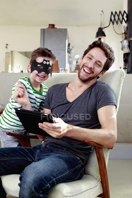 Pai e filho em máscara de morcego sentado na cadeira com tablet digital — Fotografia de Stock