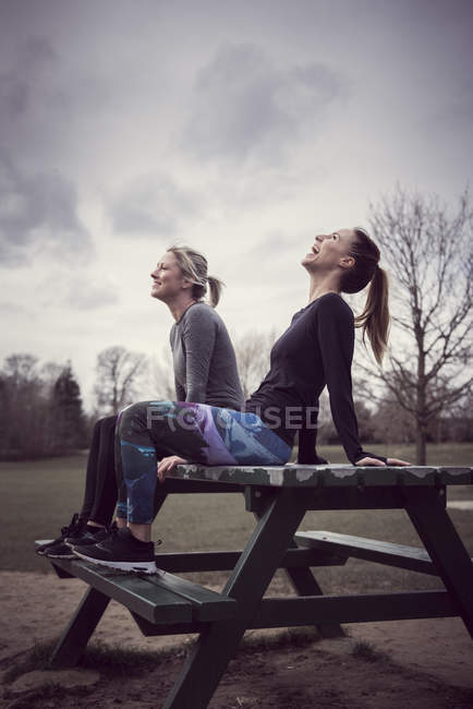 Women wearing sports clothing sitting on picnic table, head back laughing — Stock Photo