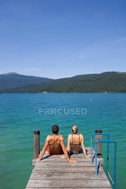Vista trasera de la pareja sentada en el muelle junto al lago - foto de stock