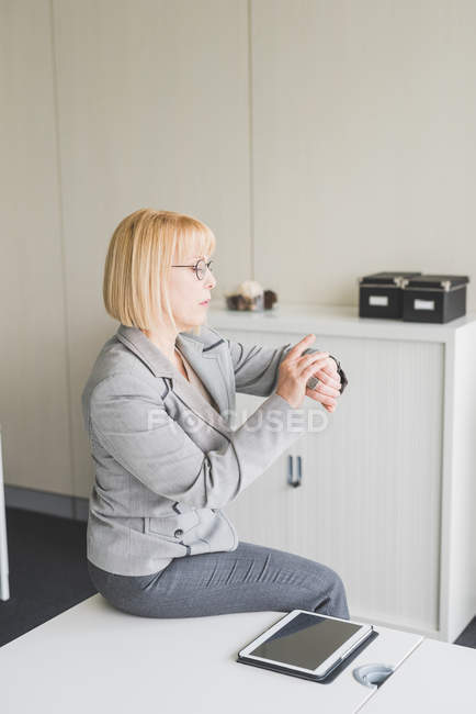 Reife Geschäftsfrau sitzt auf Büroschreibtisch und schaut auf Armbanduhr — Stockfoto
