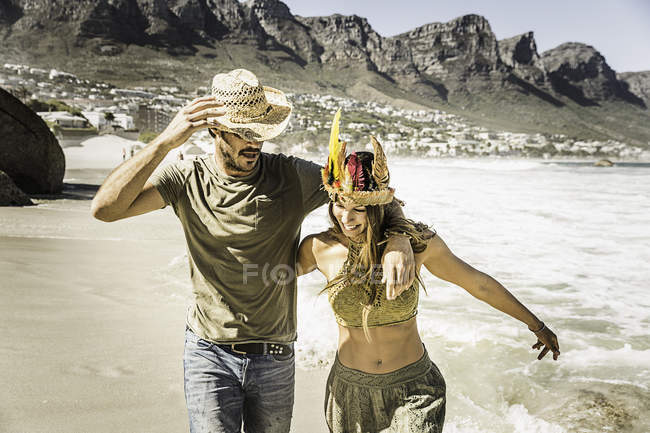 Casal adulto médio usando chapéu de palha e cobertura para a cabeça de penas correndo na praia, Cape Town, África do Sul — Fotografia de Stock