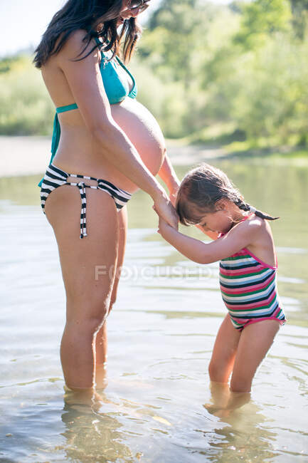Беременные мать и дочь стоят в озере, держась за руки — стоковое фото