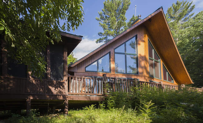 Vista trasera de una casa de estilo casa de campo fresada de tronco plano con cubierta de madera elevada y paisajismo en verano, Mt-Tremblant, Quebec, Canadá - foto de stock