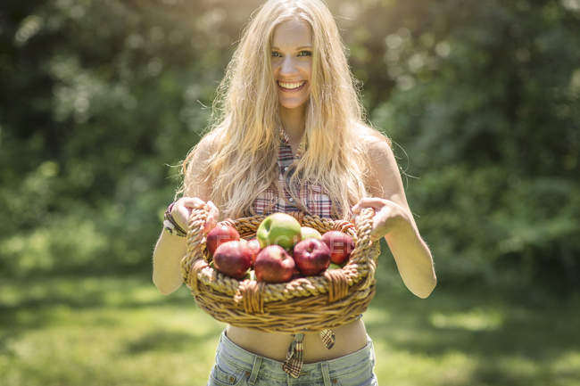 Portrait of young woman holding up basket of fresh apples in garden — Stock Photo