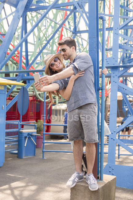 Casal contemporâneo ter um bom tempo na frente de passeio tomando selfie no parque de diversões — Fotografia de Stock