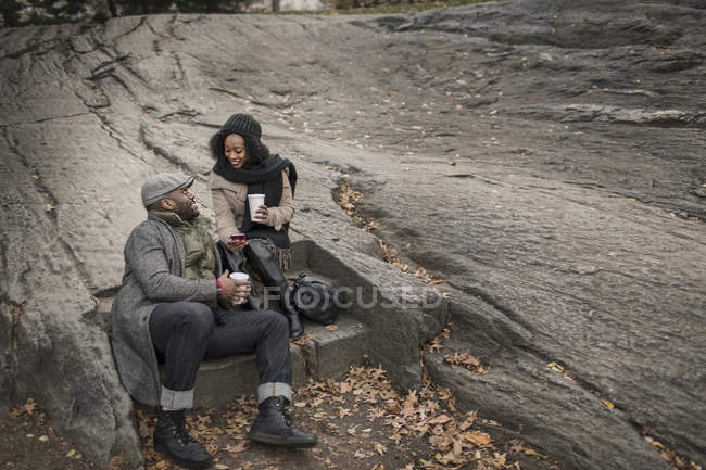 Романтическая счастливая пара наслаждается городом во время зимних каникул, пьет кофе и смотрит на смартфон в парке — стоковое фото