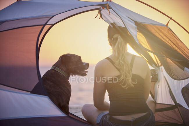 Une femme et son chien boxeur apprécient le coucher de soleil assis dans une tente de camp sur la plage. — Photo de stock