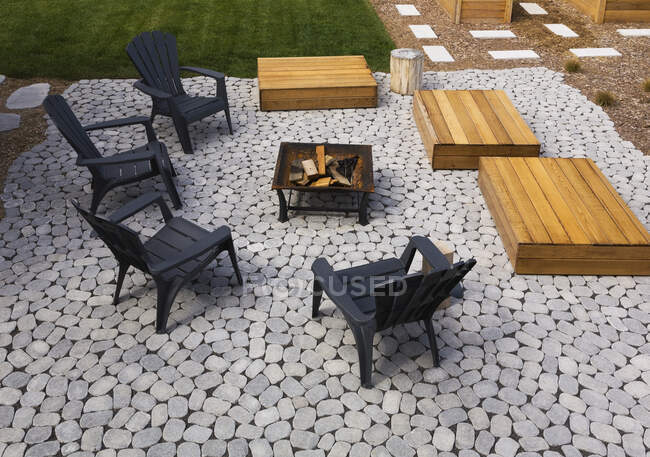 Grey paving stone patio with black Adirondack sitting chairs, slightly raised flat rectangular red cedar wooden bench platforms around a metal firepit in residential backyard in summer, Quebec, Canada — Stock Photo