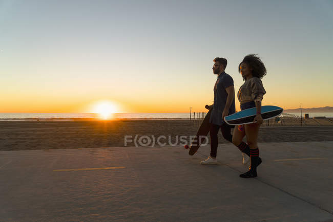 Пара, що йде уздовж доріжки на пляжі, тримає скейтборди — стокове фото