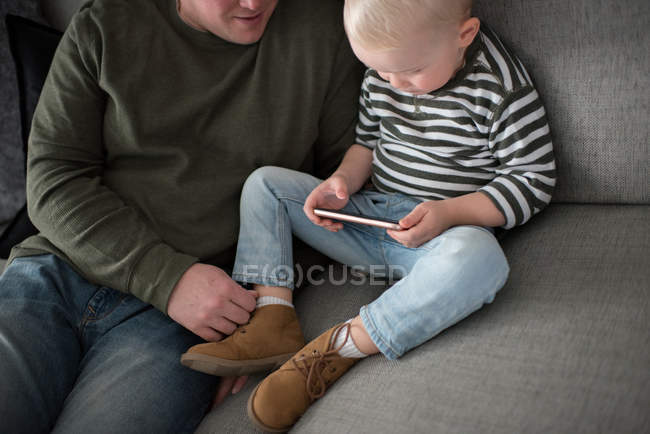 Отец и сын сидят на диване, сын смотрит на смартфон — стоковое фото