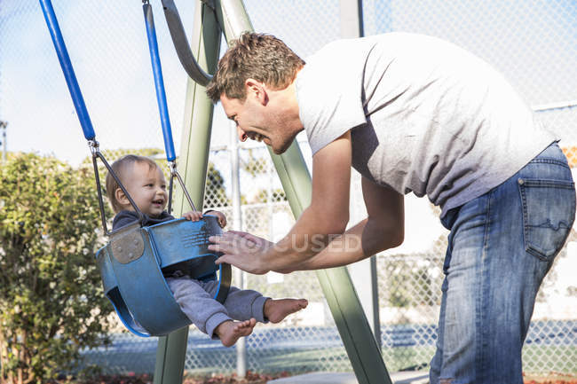 Padre spingendo giovane figlio su altalena parco giochi — Foto stock