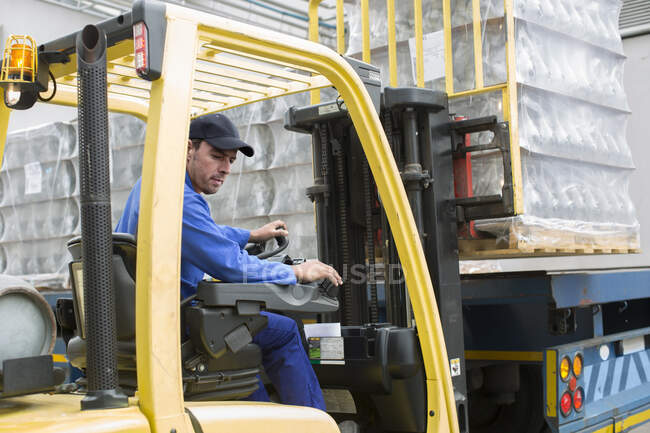 Cape Town, South Africa,male worker in overalls seated lifting up case of plastic wrapped bottles from forklift in packaging factory — Stock Photo