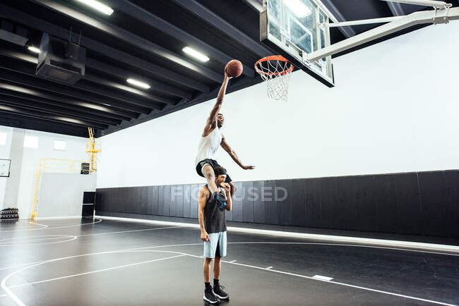 Male basketball player getting shoulder carry to throw ball in basketball hoop — Stock Photo