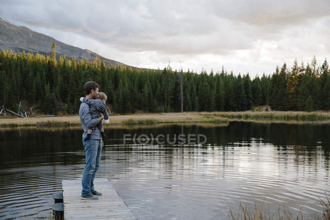 Père debout sur une jetée en bois au bord du lac, tenant un jeune fils — Photo de stock